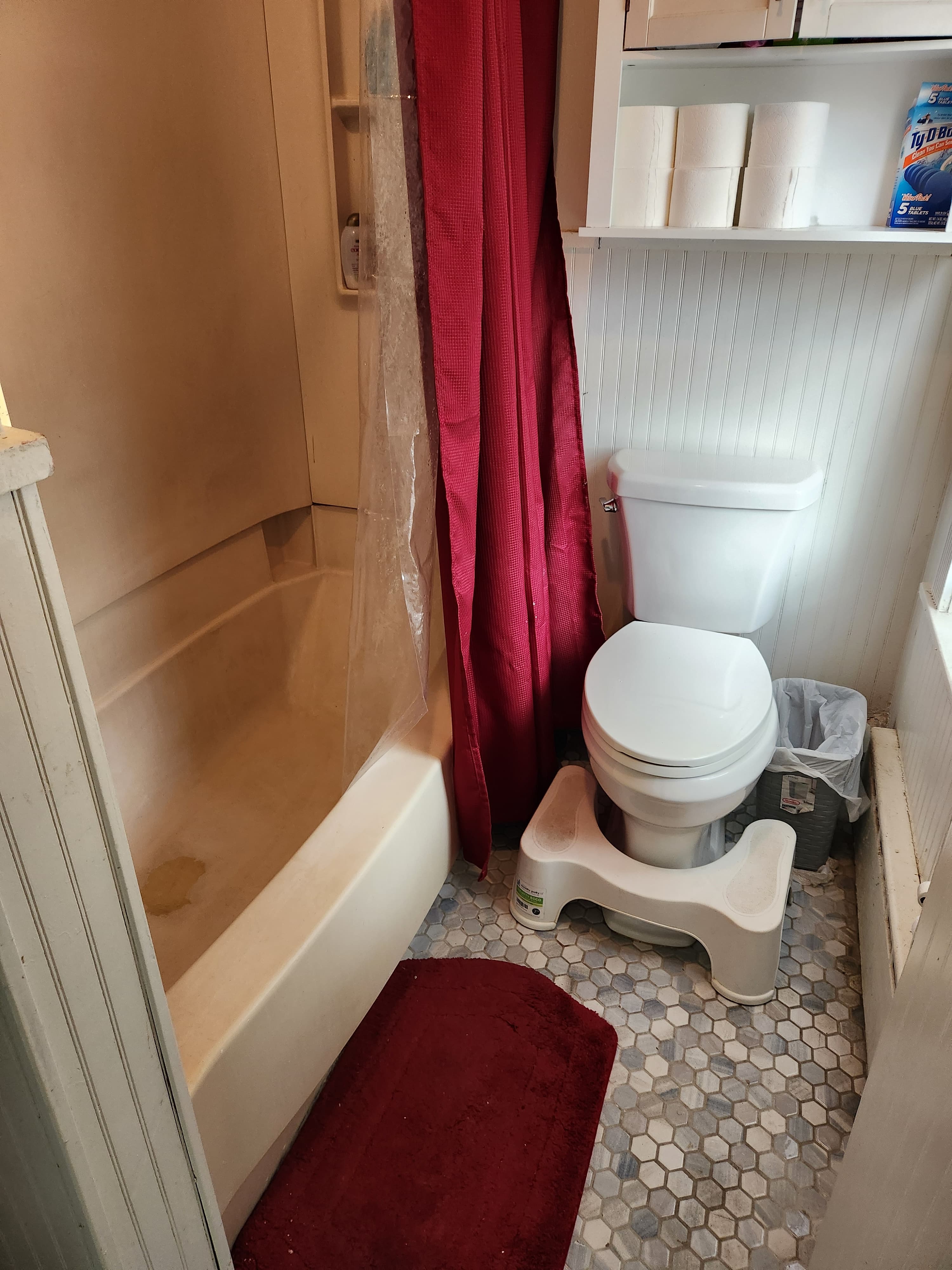 Small bathroom with a bathtub, red curtain, white toilet, stool, and hexagonal tile floor.