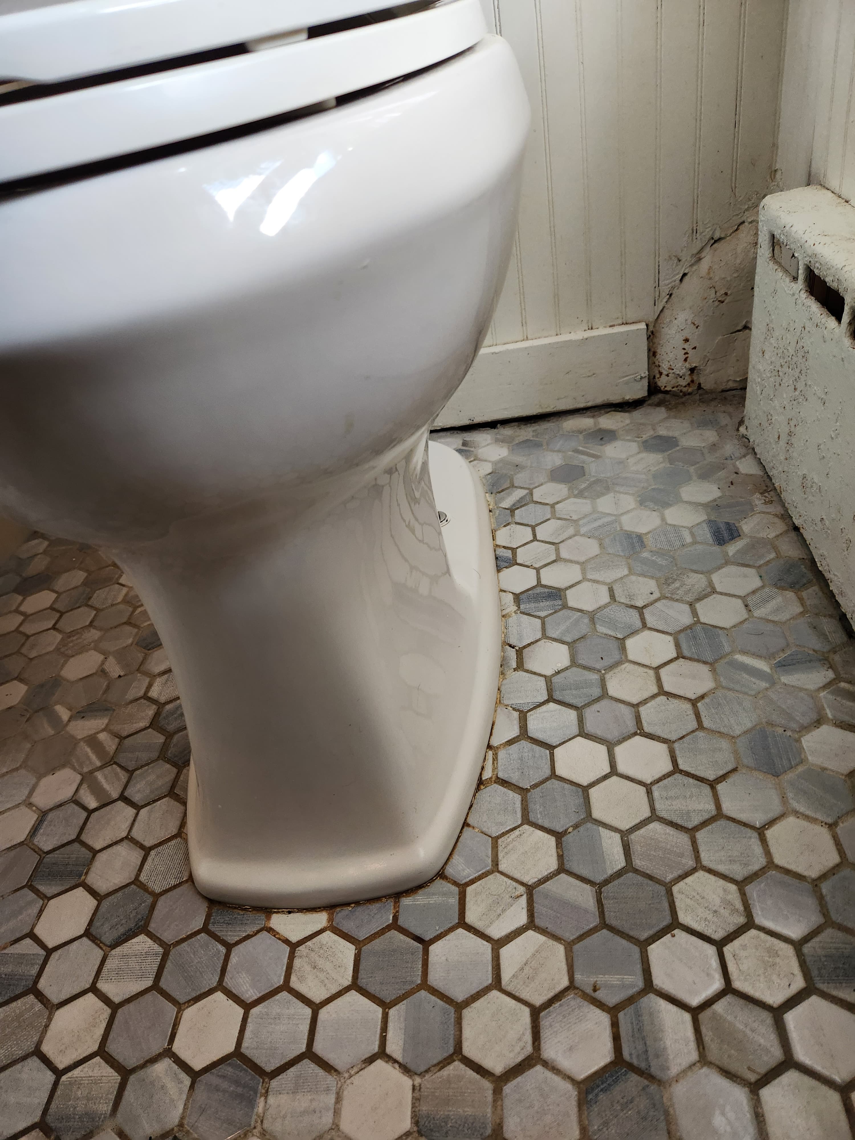 White toilet base on grey and white hexagonal tile floor beside a beadboard wall.