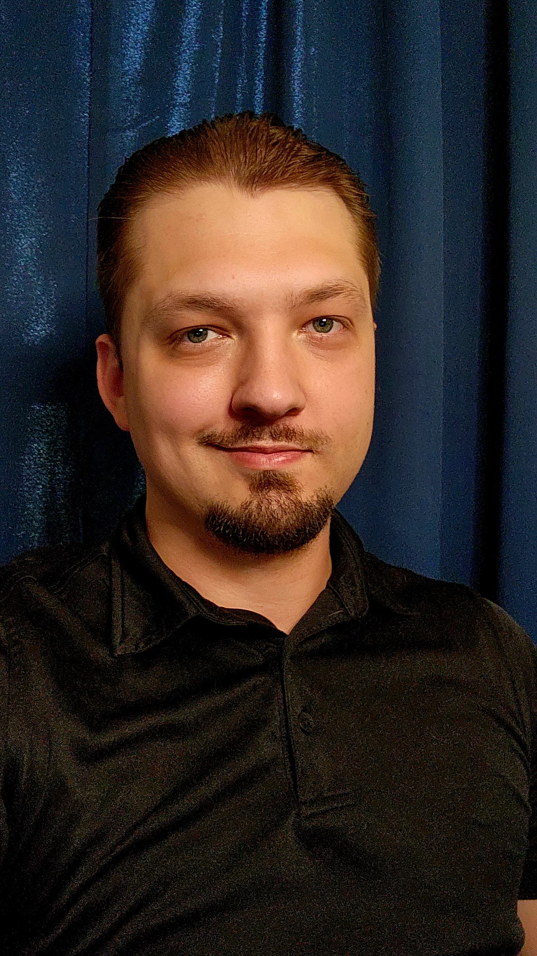 Man with slicked-back hair and a goatee wearing a black polo shirt against blue curtains.