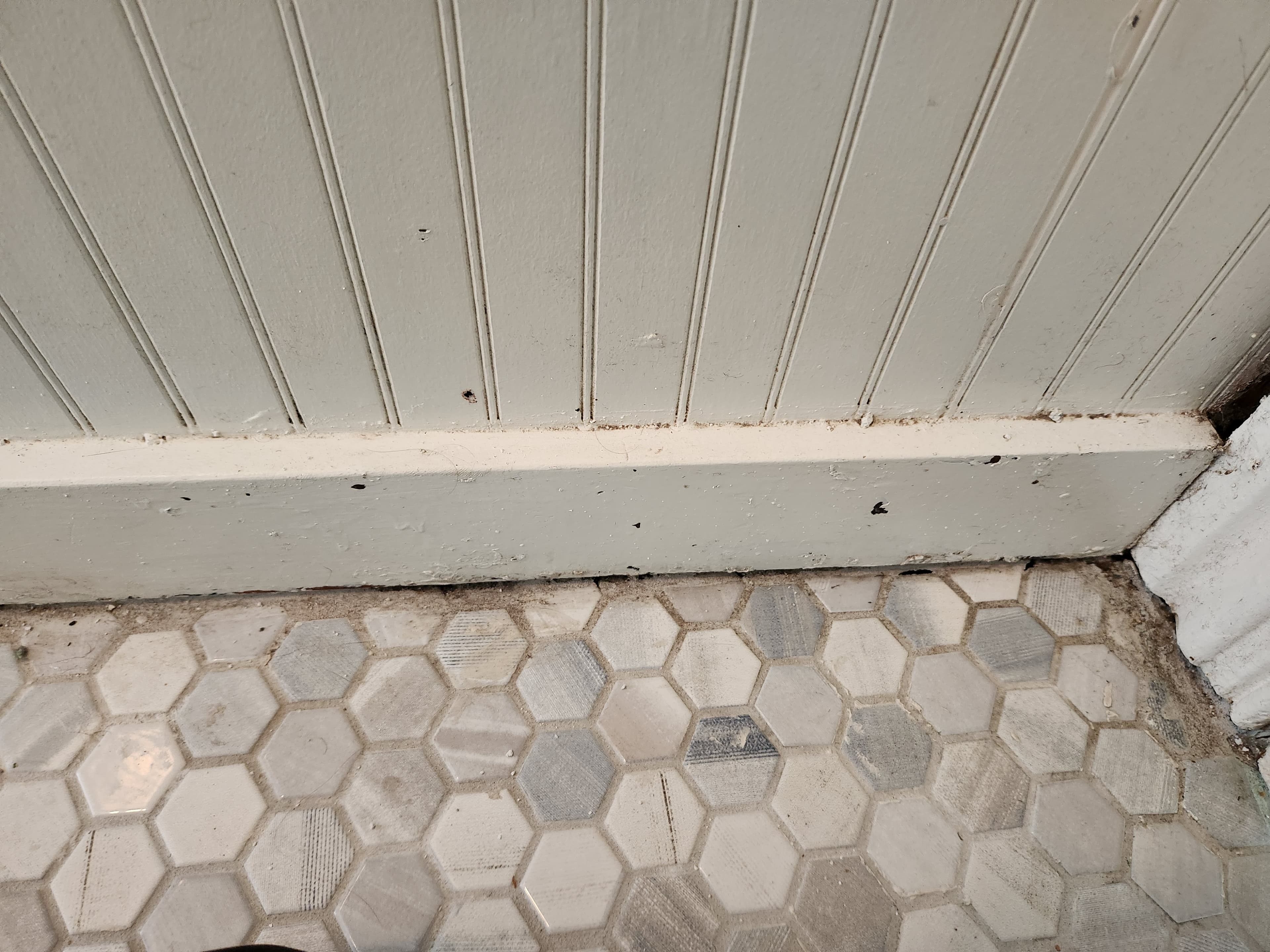 White beadboard wall and baseboard above a gray and white hexagonal tile floor with debris.